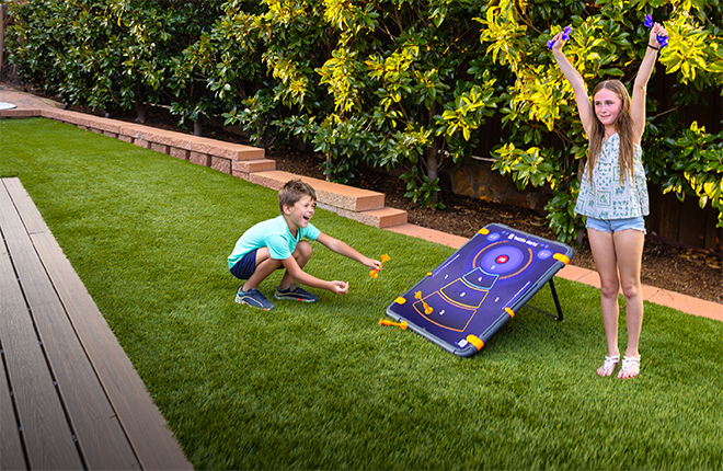 A children's outdoor target game set up on a green lawn, with a boy aiming a dart and a girl standing nearby with her arms raised. The target board is purple with a target design and yellow dart holders, set against a background of a garden and wooden deck.